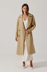 A woman is wearing a long, beige trench coat with double-breasted buttons, wide lapels, and buttoned straps at the cuffs. Underneath, she has on a white satin dress with a deep V-neckline and a slit that reveals light-colored shoes.